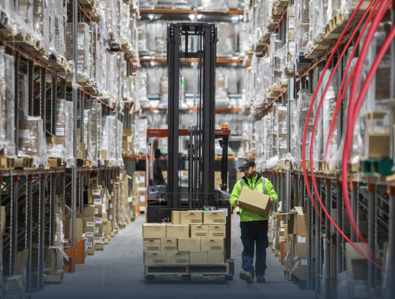 A warehouse worker in a high-visibility jacket carrying a box, with shelves stocked full of packaged goods and a forklift in operation in the background.